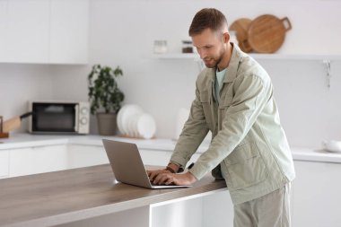 Genç sakallı adam mutfaktaki masada dizüstü bilgisayar kullanıyor.