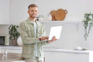 Genç sakallı adam mutfakta dizüstü bilgisayar kullanıyor.