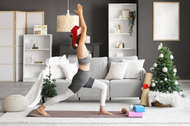 Noel Baba şapkalı sportif olgun kadın Noel arifesinde evde yoga yapıyor.