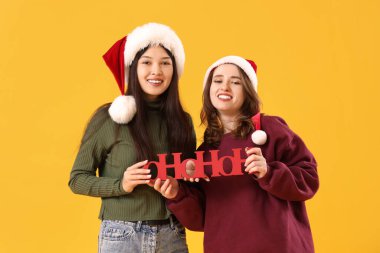 Young lesbian couple in Santa hats with Christmas greeting on yellow background clipart