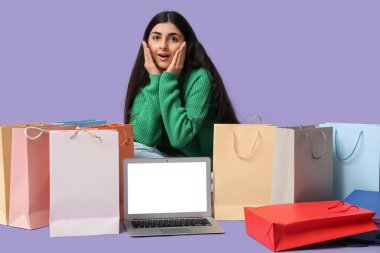 Alışveriş çantaları ve laptopuyla leylak arka planda oturan şaşırmış genç bir kadın.