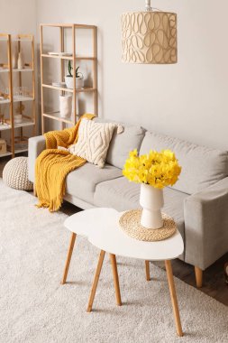 Konforlu kanepe, raf ünitesi ve oturma odasında vazo dolusu narsist çiçekli kahve masası.