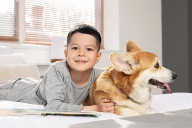 Little happy Asian boy reading book with cute Corgi dog lying at home