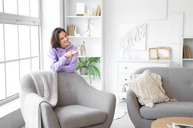 Klimalı gülümseyen genç kadın oturma odasındaki koltuğun yanında duruyor.