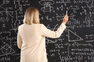 Olgun kadın matematik öğretmeni sınıfta tahtaya tebeşirle yazı yazıyor.