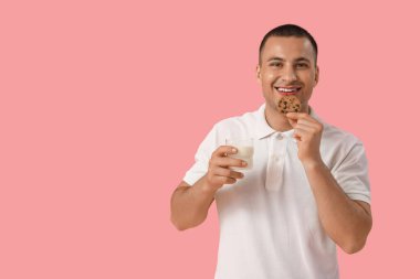 Pembe arka planda bir bardak sütle kurabiye yiyen genç adam.