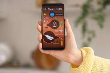 Akıllı ev güvenlik kontrol paneli olan cep telefonlu genç bir kadın mutfakta, yakın plan.