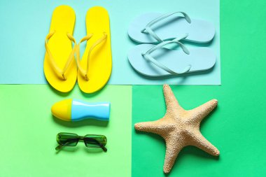 Değişik parmak arası terlikler, güneş gözlükleri, deniz yıldızı ve renk arkaplanında bir şişe güneş kremi.