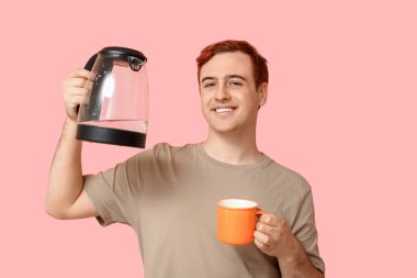 Modern elektrikli su ısıtıcısı ve pembe arka planda kupası olan yakışıklı bir genç.