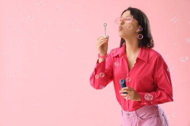 Mutlu genç bir kadın pembe arka planda sabun köpükleri üflüyor.