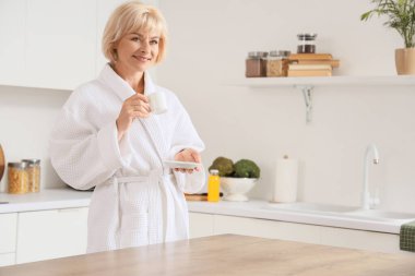 Mutfakta bir fincan kahveyle bornozlu orta yaşlı bir kadın.