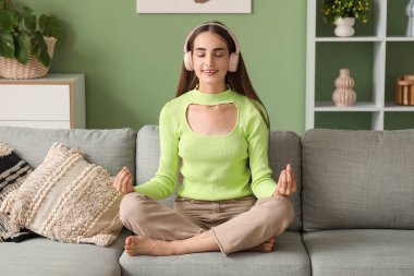 Kulaklık takan, diyabetik hastası genç bir kadın evdeki kanepede meditasyon yapıyor.