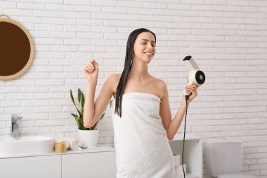 Saçında saç kurutma makinesi ve maskesiyle banyoda genç ve güzel bir kadın.