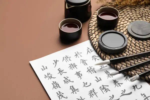 stock image Paper sheet with Asian hieroglyphs, brushes, ink and cups of tea on color background, closeup. International Haiku Poetry Day