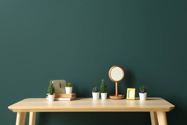 Yeşil duvarın yanındaki masa üzerinde kitap, saat ve ayna bulunan yapay bitkiler.