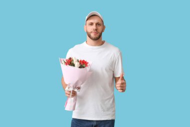 Güzel lalelerle dolu bir buketi olan genç teslimatçı mavi arka planda baş parmak hareketi gösteriyor.