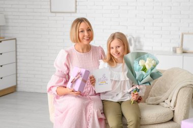 Elinde hediye kutusu olan güzel bir genç kadın ve oturma odasındaki kanepede elinde bir buket beyaz lale ve tebrik kartı olan küçük tatlı kızı.