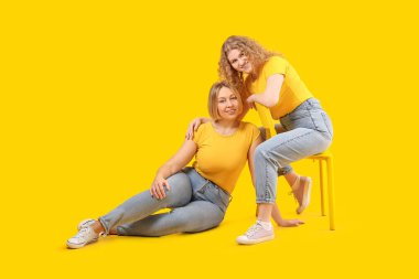 Şık kotlu güzel, mutlu kadınlar sarı arka planda sandalyede oturuyorlar.