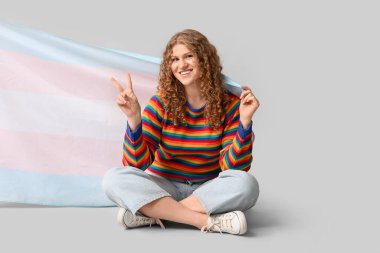 Gri arka planda oturan transseksüel bayrağı olan güzel genç bir kadın zafer jesti sergiliyor.