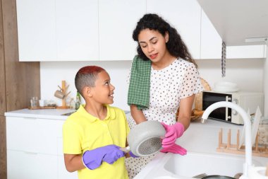 Annesi mutfakta bulaşık yıkayan Afro-Amerikalı bir çocuk.