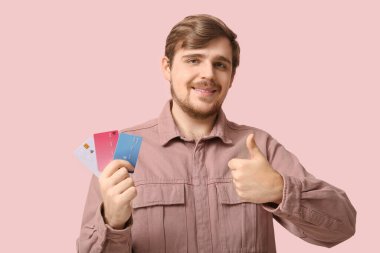 Kredi kartlarıyla gülümseyen genç adam pembe arka planda baş parmağını gösteriyor.