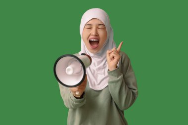 Genç Asyalı Müslüman kadın megafona bağırıyor ve yeşil arka plandaki bir şeye işaret ediyor. Ulusal Duyulma Günü