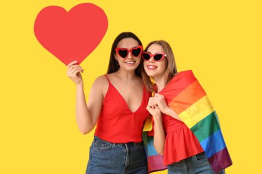 Sarı arka planda LGBT bayrağı ve kağıt kalpli güzel lezbiyen çift.