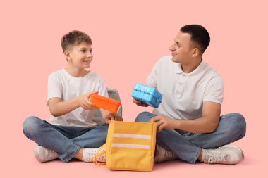 Küçük çocuk ve babası okul çantalarını pembe arka planda çantaya dolduruyorlar.