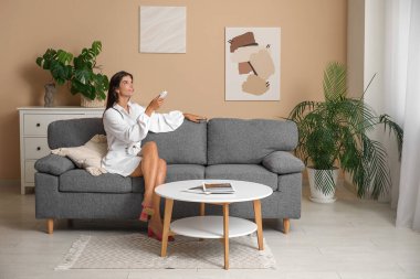 Klima kumandalı genç bir kadın oturma odasındaki bej duvarın yanındaki koltukta oturuyor.