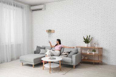 Oturma odasındaki beyaz tuğla duvarın yanındaki koltukta klimalı uzaktan kumandalı genç bir kadın.