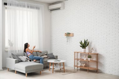 Oturma odasındaki beyaz tuğla duvarın yanındaki koltukta klimalı uzaktan kumandalı genç bir kadın.