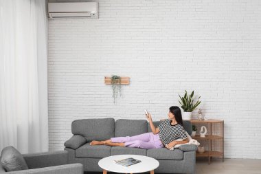 Klimalı uzaktan kumandalı genç bir kadın evdeki beyaz tuğla duvarın yanındaki kanepede yatıyor.