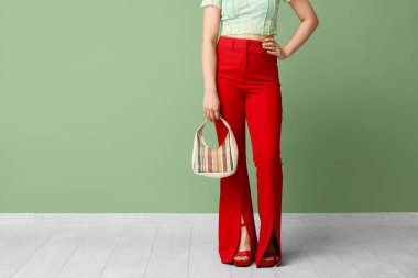 Kırmızı pantolonlu şık bir genç kadın ve yeşil duvarın yanında el çantalı topuklu ayakkabılar.