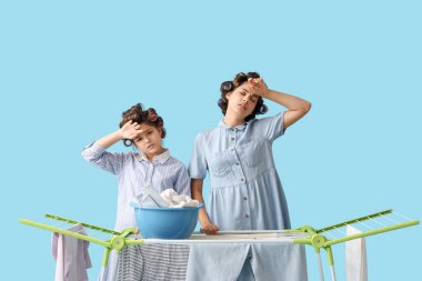 Yorgun küçük kız ve annesi saç bigudileriyle kurutucuya mavi arka planda çamaşır asıyorlar.