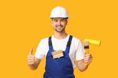 Sarı arka planda baş parmağını gösteren boya silindirli ve fırçalı erkek ressamın portresi