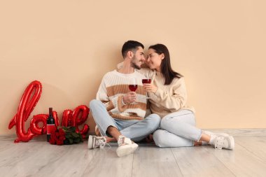 Şarap bardakları, balonları ve gülleri olan mutlu genç çift bej duvarın yanında oturuyor. Sevgililer Günü kutlaması