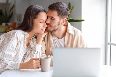 Dizüstü bilgisayarı olan mutlu genç çift mutfakta çay içiyor.