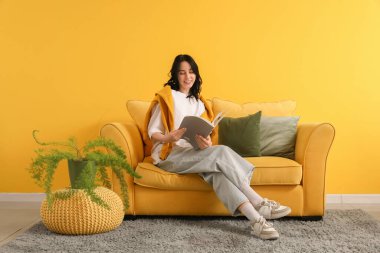 Evdeki sarı kanepede dergi okuyan güzel genç bir kadın.