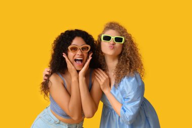 Güneş gözlüklü güzel genç kadınlar sarı arka planda öpüşüyorlar.