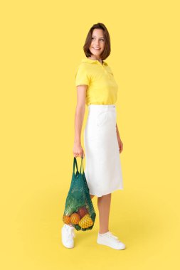 Sarı arka planda taze sebze ve meyvelerle dolu bir sicim torbası olan güzel, genç ve mutlu bir kadın. Diyet kavramı