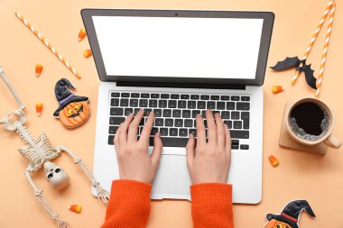 Dizüstü bilgisayarı, kahvesi ve bej arka planda Cadılar Bayramı süslemeleri olan kadın elleri.