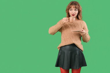 Yeşil arka planda bir fincan çayla sonbahar kıyafetleri içinde şık genç bir kadın.