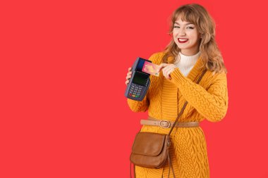 Kırmızı arka planda kredi kartı ve ödeme terminali olan şık genç bir kadın.