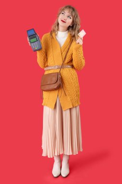 Kırmızı arka planda kredi kartı ve ödeme terminali olan şık genç bir kadın.