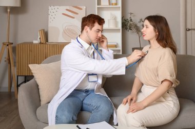 Erkek doktor evde genç bir kadının ciğerlerini dinliyor.