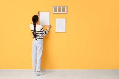 Genç bir kadın odadaki turuncu duvara resim çerçevesi asıyor.