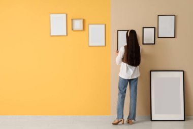 Genç bir kadın odadaki turuncu duvara resim çerçevesi asıyor.
