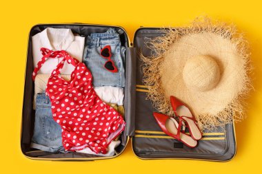 Sarı arka planda kadın kıyafetleri, şapka ve ayakkabıları olan bavulu aç. Seyahat kavramı