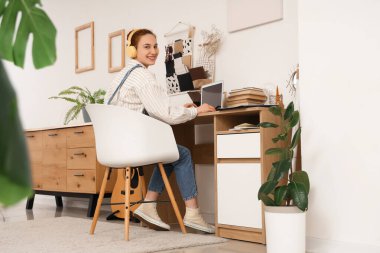 Kulaklıklı genç kız evdeki masada bilgisayar kullanıyor.