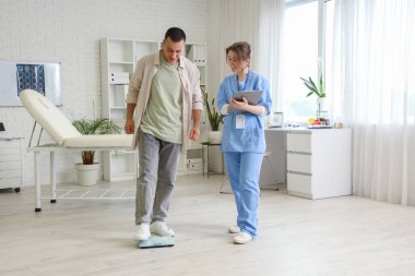 Klinikte doktorla tartıya göre ağırlığını ölçen genç bir adam.